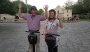Two happy people are waving while standing on Segways in front of a historical building with other visitors in the background.