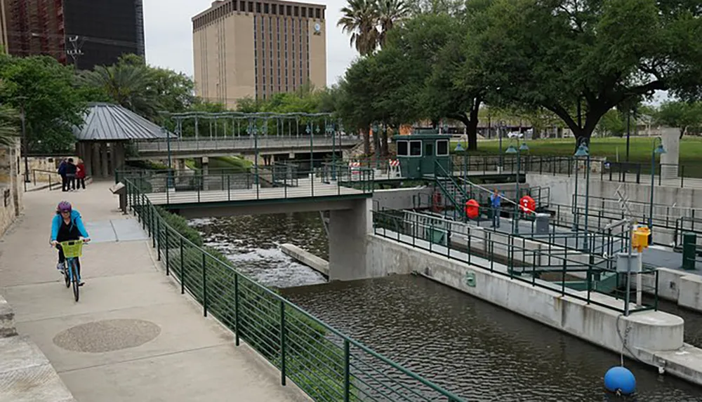 A person is cycling along a riverwalk next to an urban waterway with a bridge and gate mechanism with pedestrians and greenery in the background