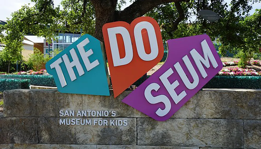 The image displays a colorful playful sign for The DoSeum which is San Antonios museum for kids positioned against a backdrop of trees flowers and a clear sky