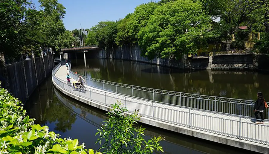 A curved pedestrian bridge spans a serene canal, flanked by green trees and a clear blue sky, as people enjoy a sunny day out.