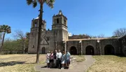 A group of people is posing for a photo in front of an old Spanish colonial church with a clear blue sky above them.