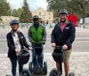 Three smiling people wearing helmets are standing with Segways in front of the Alamo on a sunny day