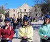 Three smiling people wearing helmets are standing with Segways in front of the Alamo on a sunny day