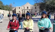 Three smiling people wearing helmets are standing with Segways in front of the Alamo on a sunny day.