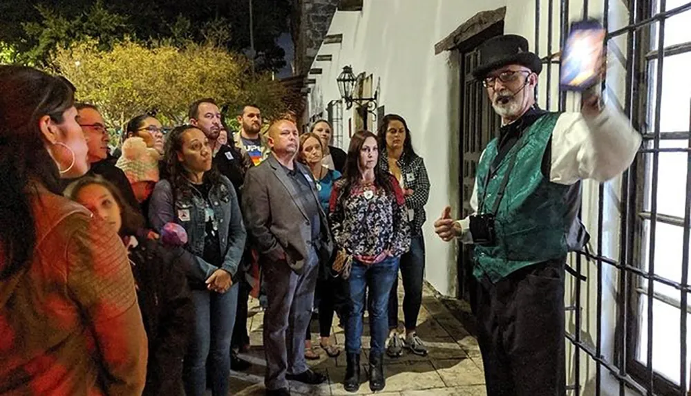 A person dressed in Victorian-style clothing is addressing a group of attentive people during what appears to be a nighttime walking tour or ghost tour