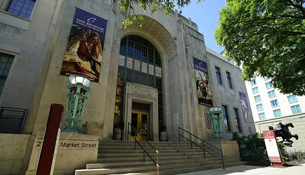 The image shows the entrance of the Briscoe Western Art Museum featuring a neoclassical facade with large arching windows decorative banners and a statue of a horse-mounted figure to the right all under a clear blue sky