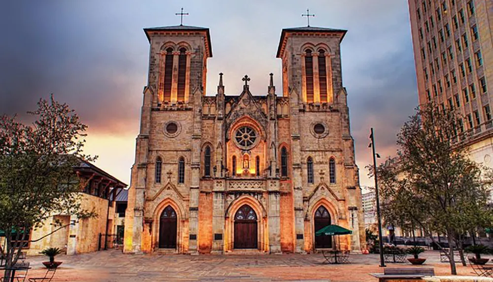 The image shows a historic cathedral with twin bell towers illuminated at dusk against a dramatic sky alongside modern urban surroundings