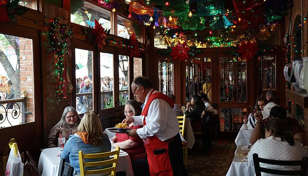 The image shows a cozy and vibrant restaurant interior with customers dining and a waiter serving food adorned with colorful festive decorations