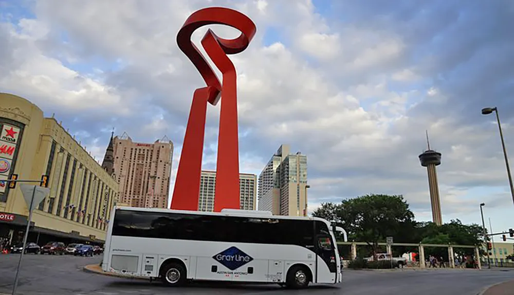 A large red sculptural torch-like structure stands prominently in an urban environment with a tour bus in the foreground and a tower in the distance under a partly cloudy sky