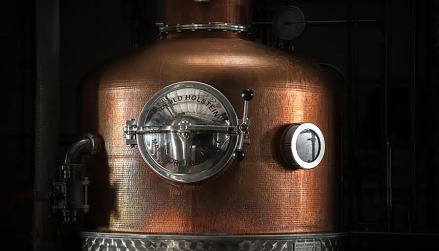 This image shows a close-up of a copper distillation apparatus, likely used for producing spirits, with a small porthole and various valves, gleaming under ambient lighting.