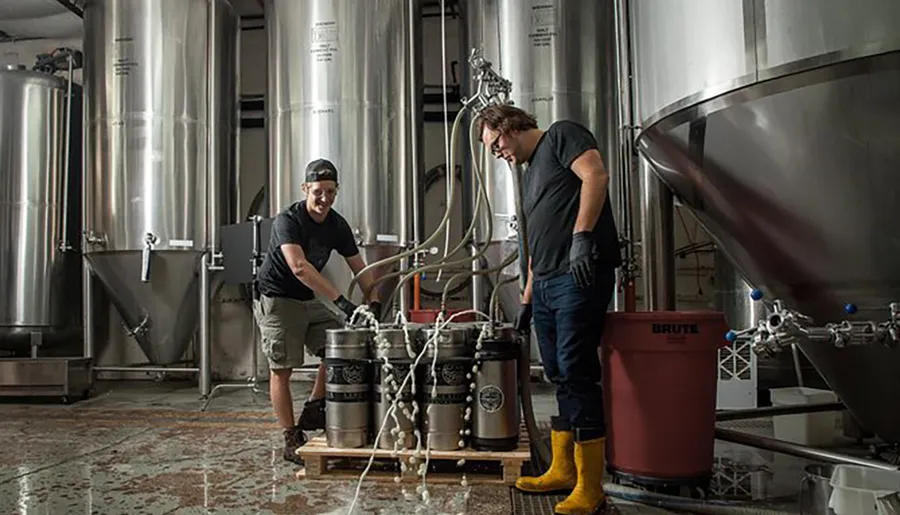 Two individuals are working with equipment and kegs in a brewery filled with large stainless steel fermenting tanks.