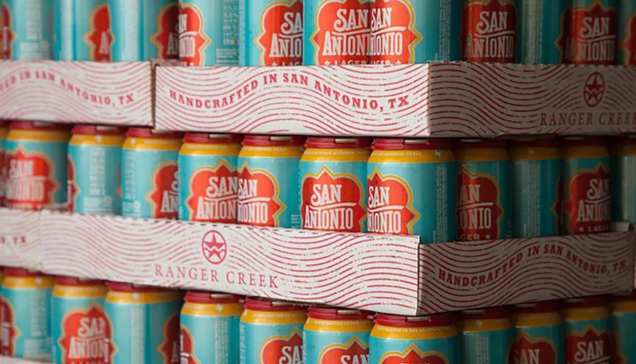 The image shows a stack of brightly colored cans of San Antonio Lager beer, handcrafted in San Antonio, TX by Ranger Creek.