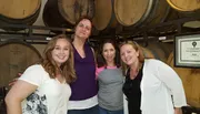 Four women are smiling for a photo in front of wooden barrels, possibly at a winery or brewery.