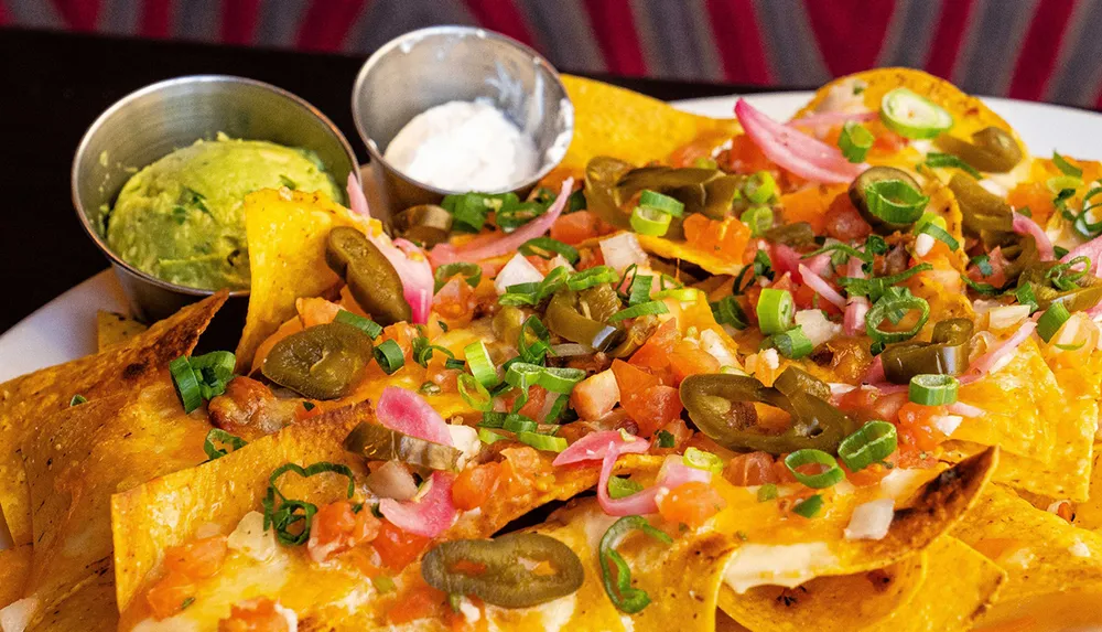 The image shows a colorful plate of nachos topped with melted cheese jalapeos tomatoes onions and green onions accompanied by sides of guacamole and sour cream