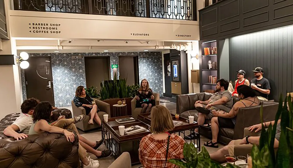 A group of people relaxes and converses in a stylish lobby area with signs indicating a barbershop restrooms coffee shop and directory
