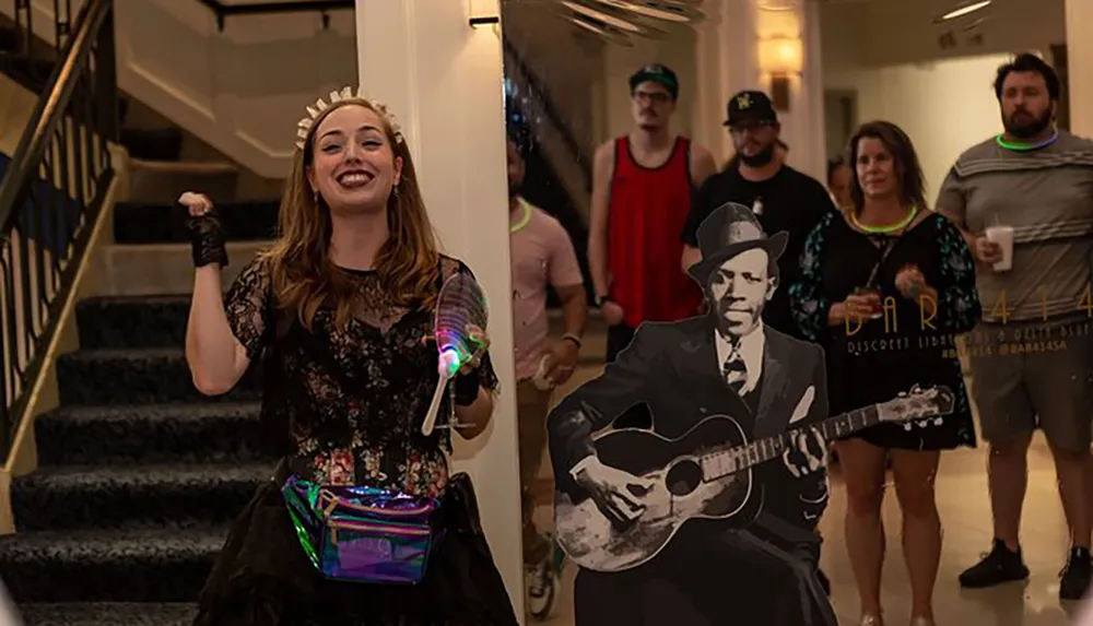 A joyous woman holding a light-up toy poses at the bottom of a staircase while a group of people and a cardboard cutout of a man with a guitar stand in the background