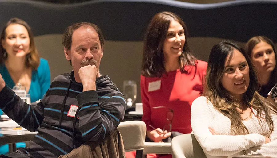 A group of people, some smiling and one appearing pensive, are attending what looks to be an indoor event or meeting.