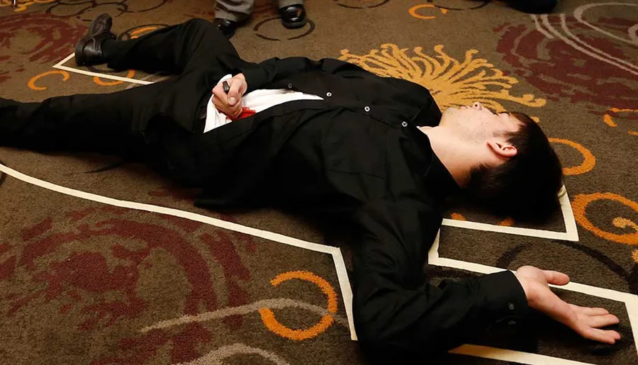 A man in a black suit is lying on the floor with his eyes closed, holding what looks like a pen and a red object in his hand, which could be part of a staged scene or simulation.