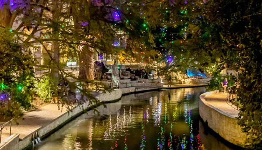 This image shows a serene nighttime scene of a tree-lined riverwalk adorned with colorful lights reflecting on the water, with a person standing by the railing enjoying the view.