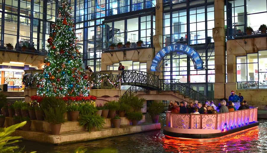 A festive scene with a decorated Christmas tree and people enjoying a boat ride along a river at night, against the backdrop of a building with the sign Rivercenter.