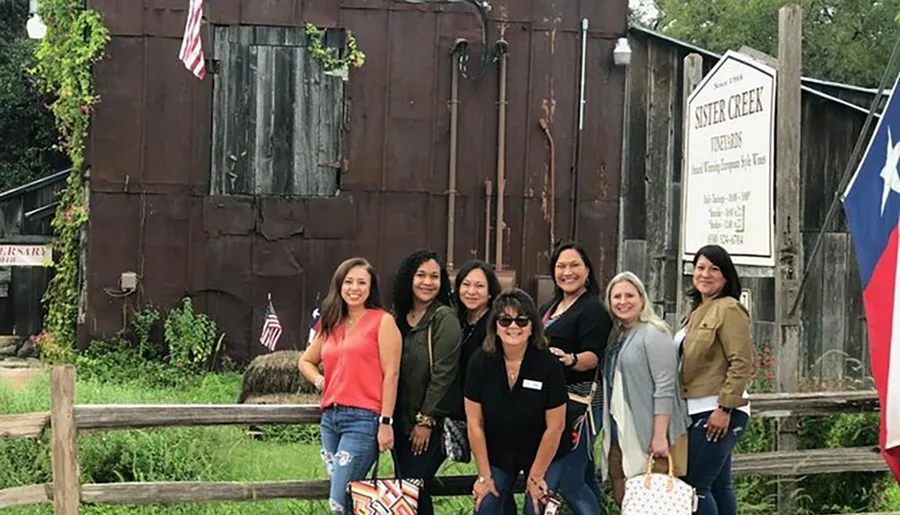 A group of people is smiling for a photo in front of a rustic building with an American flag and a sign for Sister Creek Vineyards.