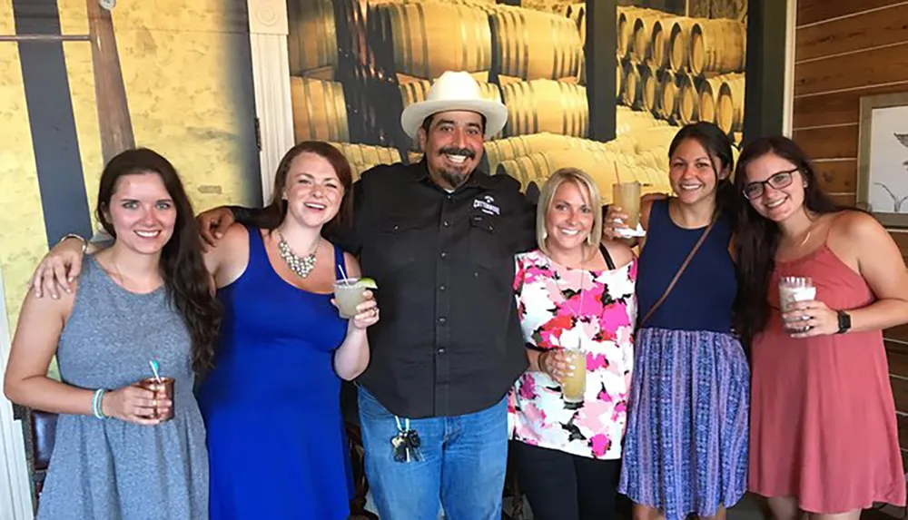 A group of five women and one man are smiling for a photo with some holding drinks in a room with a rustic decor and barrel-themed artwork on the wall