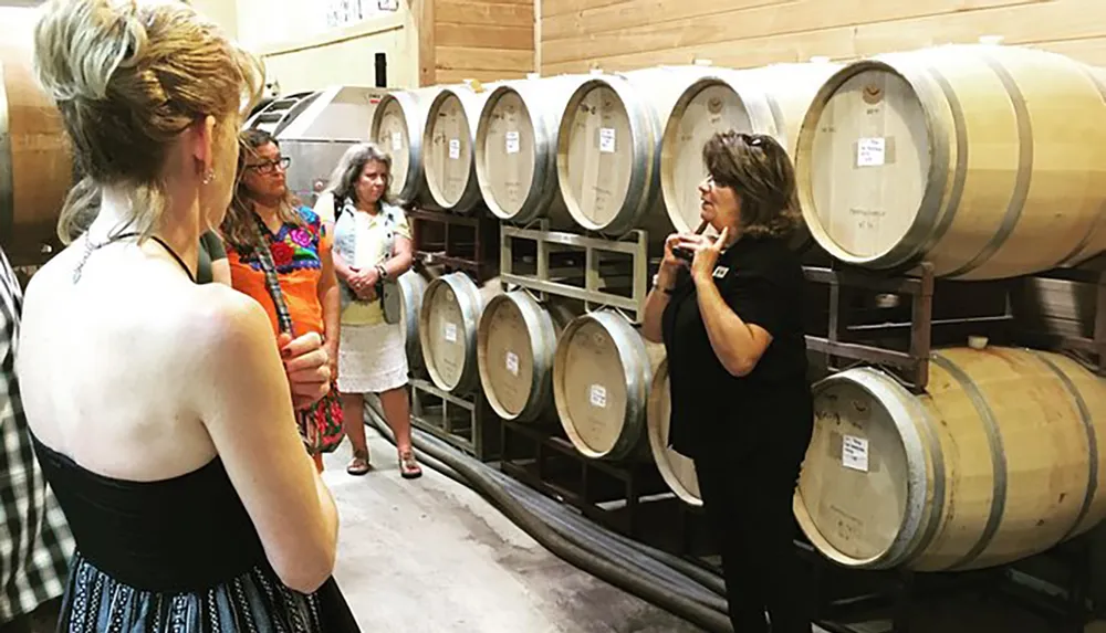 A group of individuals are attentively listening to a woman who appears to be giving a tour or presentation inside a room filled with wooden wine barrels
