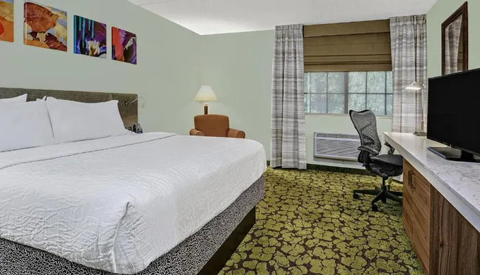A modern hotel room featuring a neatly made bed colorful artwork on the wall patterned carpet a desk with an office chair and a window air conditioning unit beneath a closed window