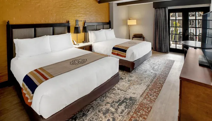The image shows a modern and neatly arranged hotel room with two beds a decorative rug a flat-screen TV and a balcony accessible through glass doors