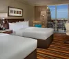 The image shows a modern well-appointed hotel room with two beds sleek furniture and a balcony that offers a city view