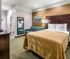 The image shows a neatly arranged hotel room with a large bed a flat-screen TV a seating area and an open door leading to the bathroom