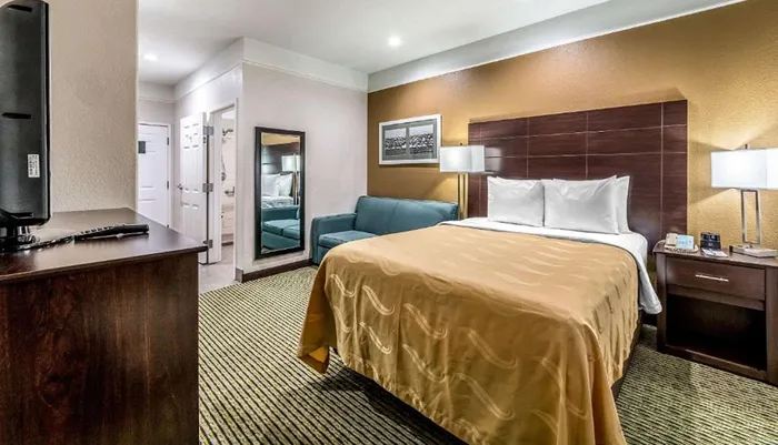 The image shows a neatly arranged hotel room with a large bed a flat-screen TV a seating area and an open door leading to the bathroom