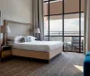 The image shows a modern and neatly arranged hotel room with a large bed a floor-to-ceiling window that offers an expansive view and contemporary furnishings