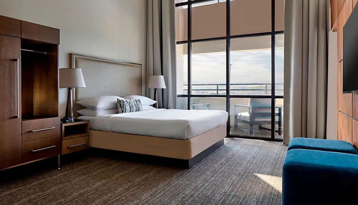 The image shows a modern and neatly arranged hotel room with a large bed a floor-to-ceiling window that offers an expansive view and contemporary furnishings