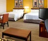 The image shows a modern hotel room with two beds a work desk with a chair a television and warm inviting decor