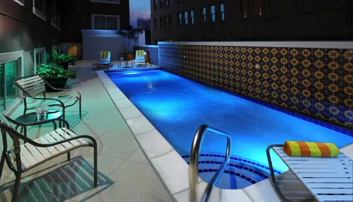 This image shows an illuminated outdoor swimming pool at nighttime with stylish poolside furniture and decorative tiles set against an urban building backdrop