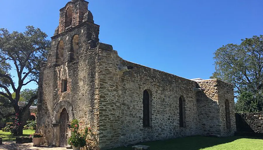 The image shows a well-preserved, historic stone mission with an ornate bell tower under a clear blue sky.