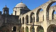 The image shows the historical architecture of an old stone mission with a dome and arches under a clear blue sky.