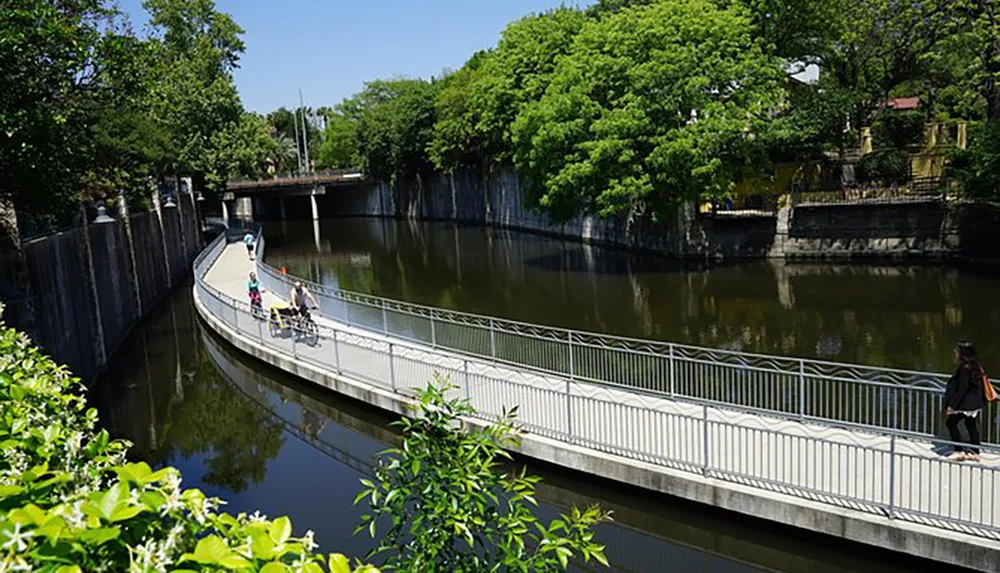 The image shows a curved pedestrian bridge over a calm canal lined with green trees under a clear blue sky with people walking and cycling across