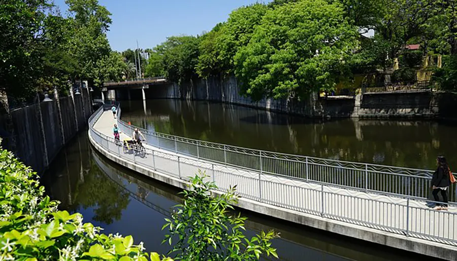 The image shows a curved pedestrian bridge over a calm canal lined with green trees under a clear blue sky, with people walking and cycling across.