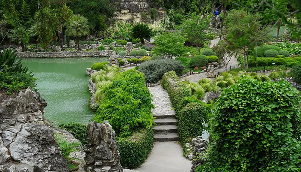The image showcases a tranquil garden with a stone pathway leading through lush greenery and ornamental shrubs alongside a serene pond