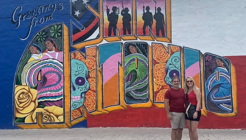 A couple is posing in front of a vibrant and colorful mural with the text Greetings from incorporated into the design