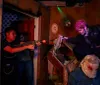 A group of people appear terrified and scream while gathered around a table with a fake gory figure as part of what looks like a haunted house or horror-themed attraction