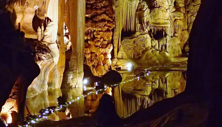 The image shows a luminous underground cave with intricate stalactites and stalagmites, reflecting beautifully in a still pool of water.