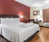 A neatly arranged hotel room with a queen-sized bed a red accent wall and a small sitting area next to the window