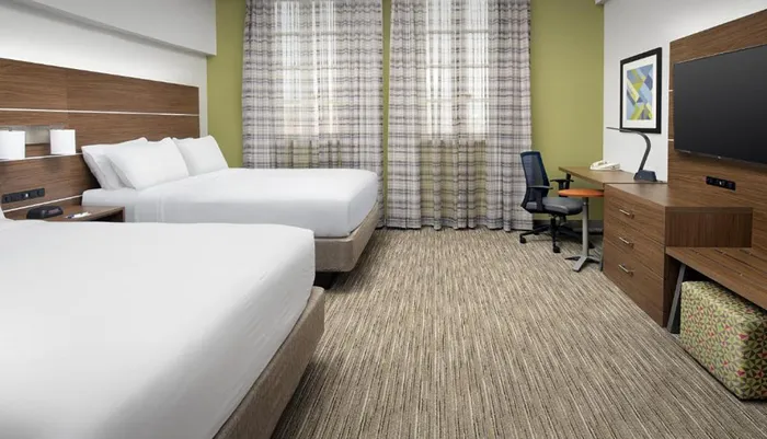 The image shows a modern hotel room with two beds a work desk with a chair a flat-screen TV and a patterned curtain covering a window
