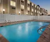 This is an image of an outdoor swimming pool enclosed by a white fence with apartment buildings in the background taken in the evening as the lights are on