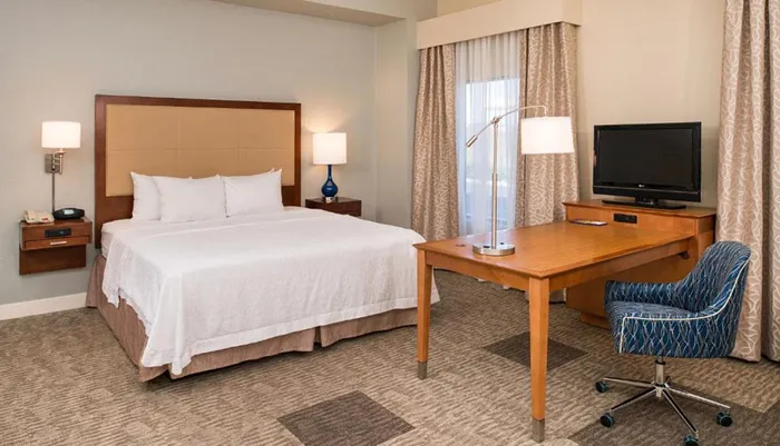 This is a neatly arranged hotel room featuring a king-sized bed a desk with a chair a television and lamps for lighting
