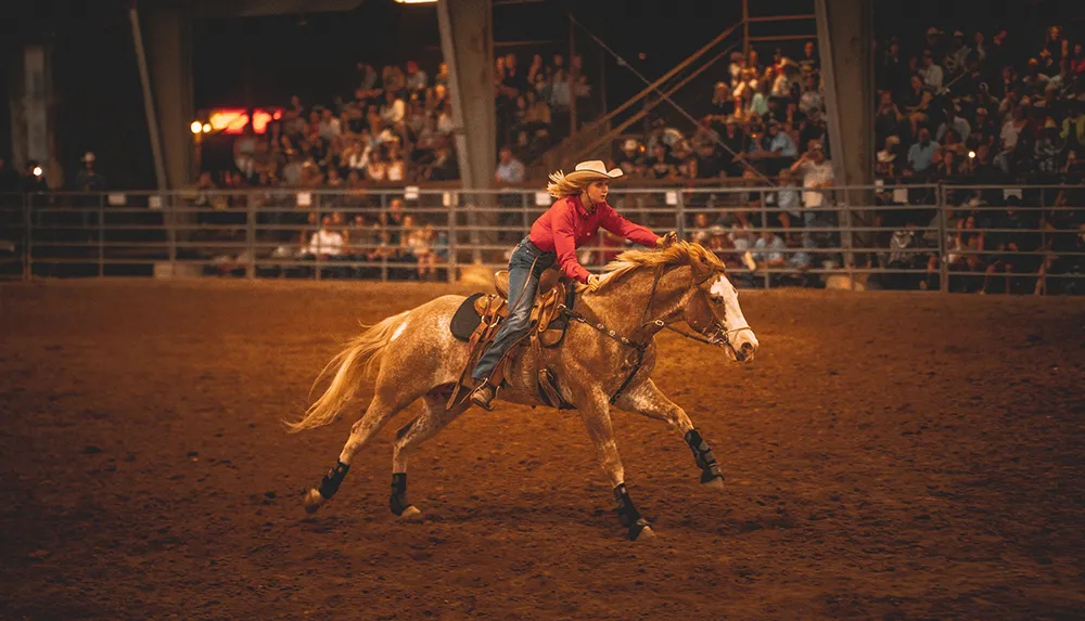 A person in a cowboy hat is riding a horse at a rodeo event with a crowd watching in the background