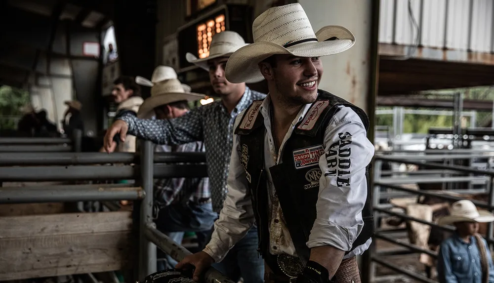 A man in a cowboy hat and rodeo gear smiles while leaning on a fence with others in similar attire in the background